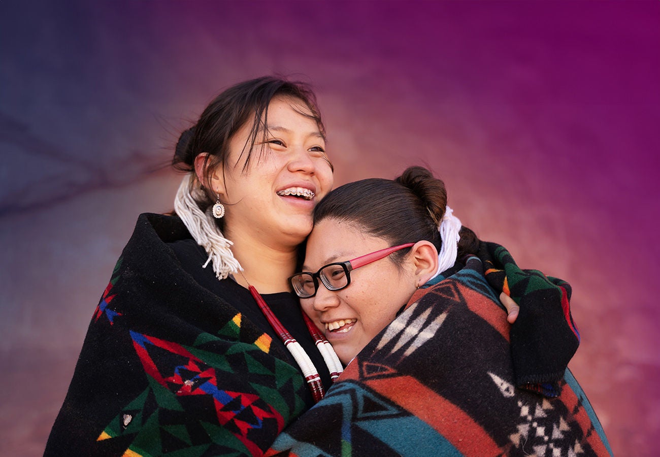 story-vibrant-fpo-women-01a-1300x900 Two women hug while wearing Native American clothing.